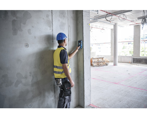 Ein Bauarbeiter mit Helm und Sicherheitsweste benutzt einen Entfernungsmesser an einer Betonwand auf einer Baustelle.