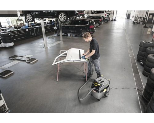 Werkstatt mit Mann beim Schleifen eines Autoteils mit Schleifgerät und Staubsauger