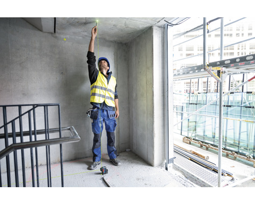 Ein Mann mit Schutzhelm und Warnweste verwendet einen Punktlaser zur Ausrichtung in einem Rohbau.