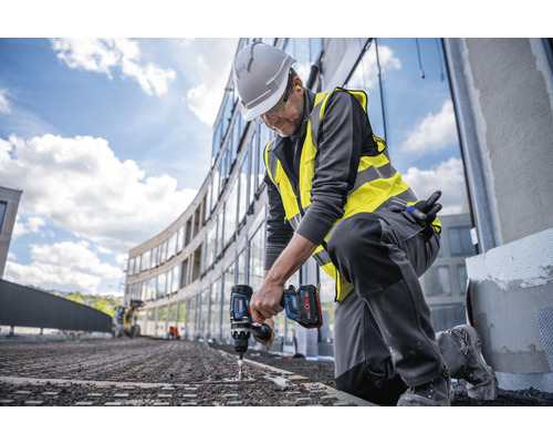 Ein Bauarbeiter kniet und verwendet einen Akku-Bohrschrauber auf einer Baustelle.