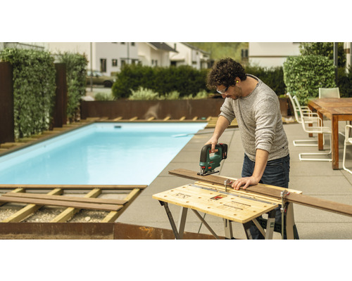 Mann schneidet Holzdiele mit einer Stichsäge im Garten, im Hintergrund ein Pool