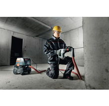 Mann mit Schutzhelm, Schutzbrille und Metabo Bohrhammer beim Bohren in eine Betonwand. Ein Staubsauger der Marke Metabo steht daneben.