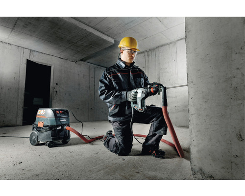 Mann mit Schutzhelm, Schutzbrille und Metabo Bohrhammer beim Bohren in eine Betonwand. Ein Staubsauger der Marke Metabo steht daneben.