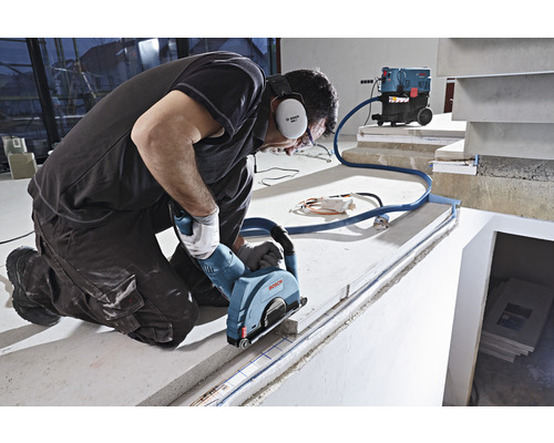 Ein Mann kniet und schneidet mit einer Mauernutfräse eine Fuge in eine Betonfläche, wobei ein Staubsauger den Staub absaugt.