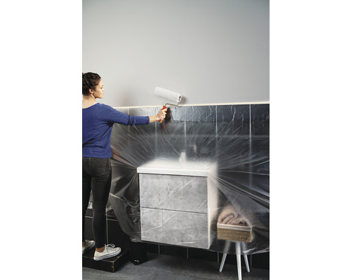 Frau streicht eine Wand mit einer Farbwalze in einem Badezimmer, das mit Folie abgedeckt ist