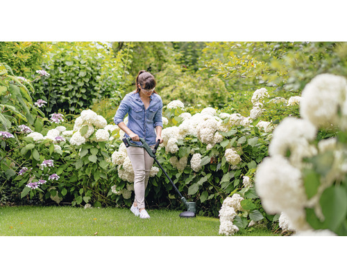 Eine Frau benutzt einen Rasentrimmer in einem Garten mit blühenden Sträuchern.