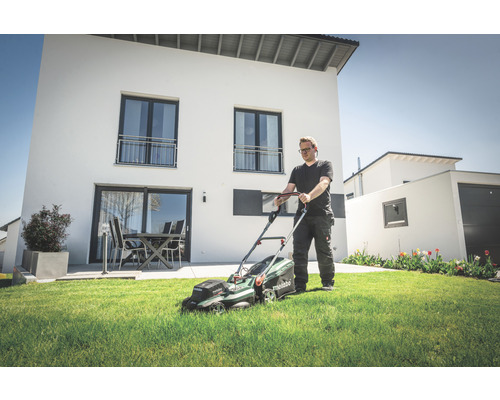Mann mäht Rasen mit einem Akku-Rasenmäher im Garten vor einem modernen Haus