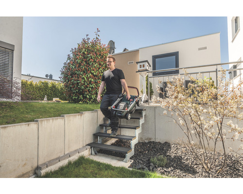 Mann trägt einen zusammengeklappten Rasenmäher eine Treppe in einem Garten hinunter