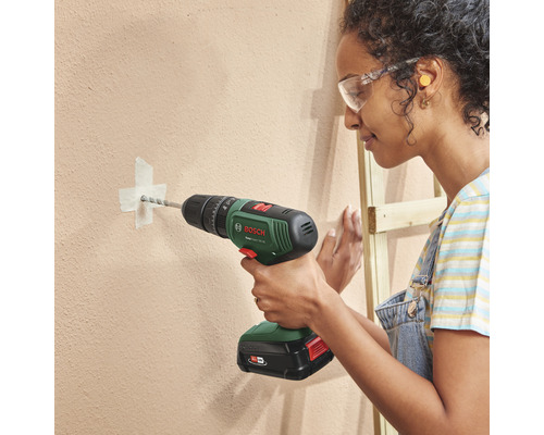 Eine Frau bohrt mit einem Akku-Bohrschrauber ein Loch in eine Wand. Sie trägt dabei eine Schutzbrille.