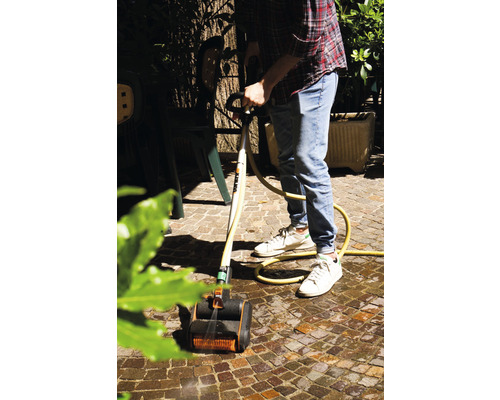 Mann reinigt Pflastersteine mit einem Bürstenreiniger und Wasserschlauch im Garten.