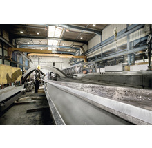 Ein Arbeiter in einer Fabrikhalle bearbeitet ein Stahlprofil mit einem Schleifwerkzeug. Im Hintergrund sind weitere Stahlkonstruktionen und ein Kran zu sehen.
