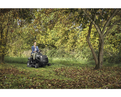Frau fährt Rasentraktor in einem Garten mit Bäumen.