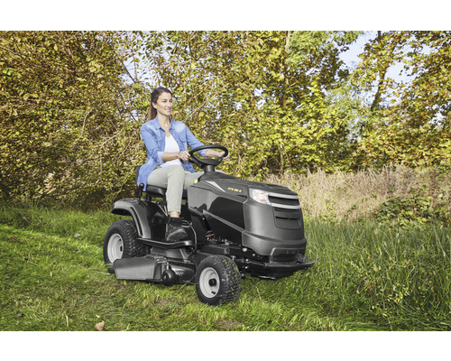 Frau fährt Rasentraktor auf Rasenfläche mit Bäumen im Hintergrund