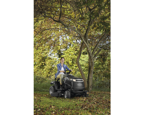 Frau fährt Rasentraktor in einem Garten mit Bäumen.