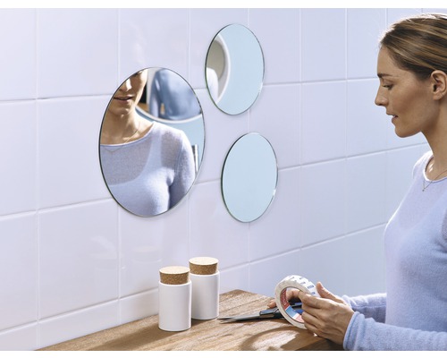 Frau befestigt runde Spiegel mit doppelseitigem Klebeband an der Wand, neben zwei weißen Behältern mit Korkdeckel