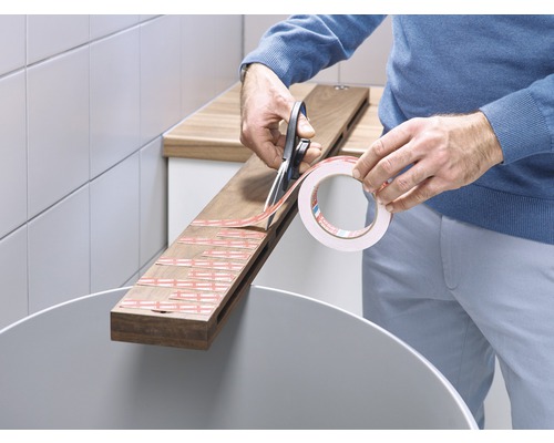 Eine Person schneidet doppelseitiges Klebeband mit einer Schere auf einem Holzbrett.