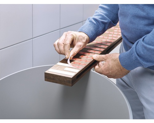 Eine Person entfernt die Schutzfolie von einem Klebestreifen auf einer Holzleiste über einem Abfalleimer.