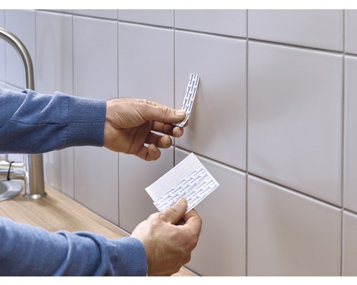 Eine Person befestigt doppelseitige Klebestreifen an einer gefliesten Wand.