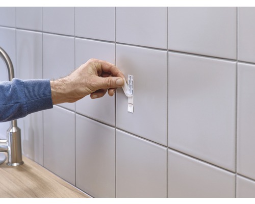 Eine Person befestigt einen Klebestreifen an einer gefliesten Wand.
