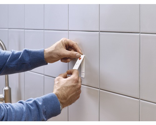 Eine Person befestigt mit einem Klebestreifen einen Gegenstand an einer gefliesten Wand.