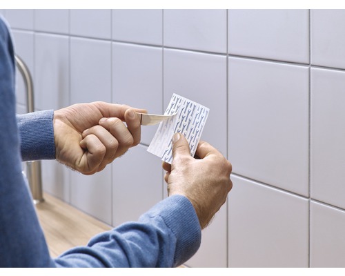 Eine Person entfernt die Schutzfolie von einem Klebestreifen vor einer gefliesten Wand.