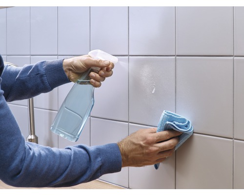 Eine Person reinigt Fliesen mit einem Sprühreiniger und einem Tuch.