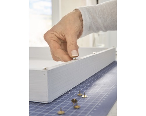 Eine Hand befestigt eine Reißzwecke an einem weißen Holzrahmen.