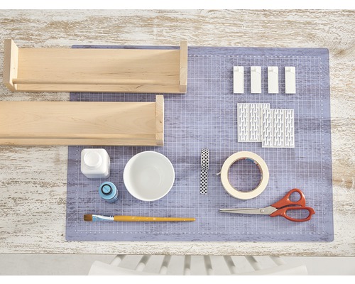 Zwei Holzregale, Farbe, Pinsel, Schere, Klebeband und Dekorpapier liegen auf einer Schneidematte.