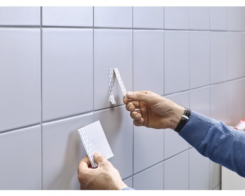 Mann befestigt mit doppelseitigem Klebeband ein Objekt an einer gefliesten Wand.