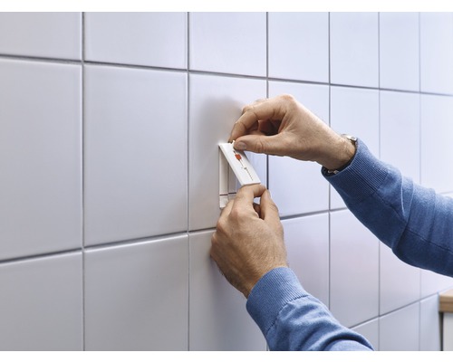 Person befestigt einen selbstklebenden Haken an einer gefliesten Wand