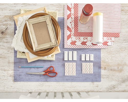 Dekorative Szene mit Bilderrahmen, Bastelpapier, Klebestreifen, Schere und Bleistift auf einer Schneidematte