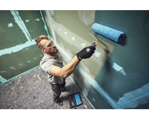 Ein Handwerker streicht eine Wand mit einer Farbrolle in einem Raum mit vorbereiteten Gipskartonplatten.