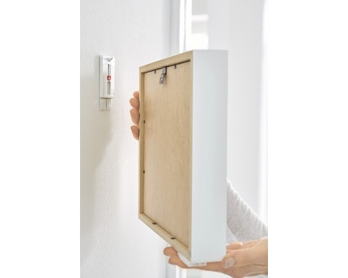 Eine Person befestigt einen Bilderrahmen mit einem Klebenagel an einer Wand.