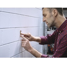 Ein Mann befestigt ein Wandregal mit einem Nagel an einer Wand aus Betonsteinen.
