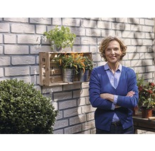 Frau steht vor einer Wand mit Verblendsteinen und einem Holzkasten mit Pflanzen.