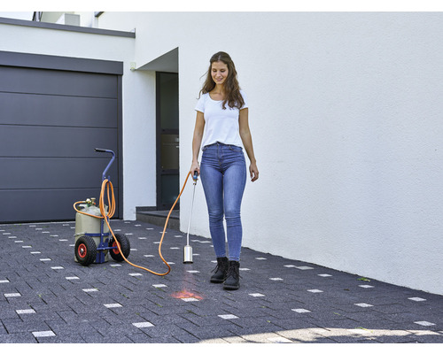 Frau verwendet einen Gasbrenner zur Unkrautbekämpfung auf einer gepflasterten Auffahrt vor einem Haus mit Garage