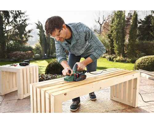 Mann hobelt Holzbank mit Elektrohobel im Garten.