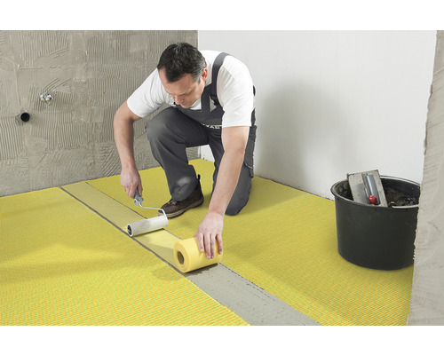 Ein Handwerker bringt mit einer Farbwalze Entkopplungsplatten auf einem Boden an. Ein Eimer mit Werkzeug steht daneben.