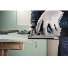 Nahaufnahme einer Stichsäge beim Schneiden einer Gipskartonplatte, bedient von einer Person mit Handschuhen.