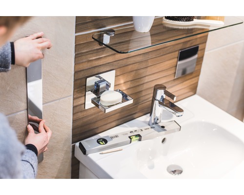 Helles Badezimmer mit Waschbecken, Seifenschale, Glasablage und Person, die einen Spiegel anbringt