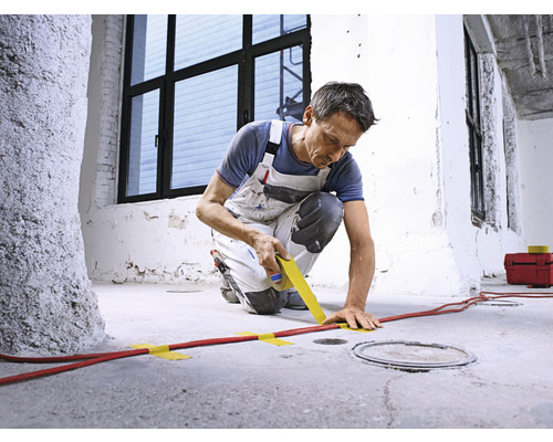 Ein Mann fixiert mit Klebeband eine rote Leitung auf einem Betonboden.