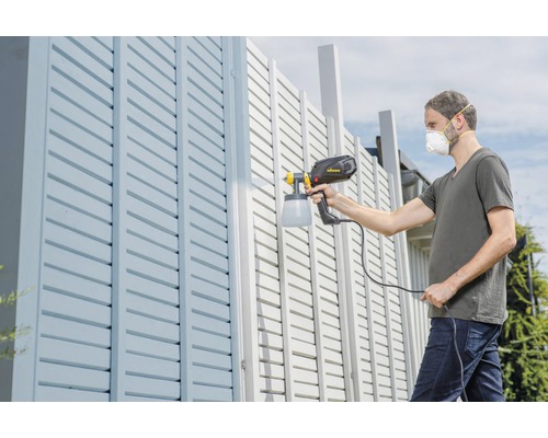 Mann streicht mit Farbsprühgerät einen Zaun.