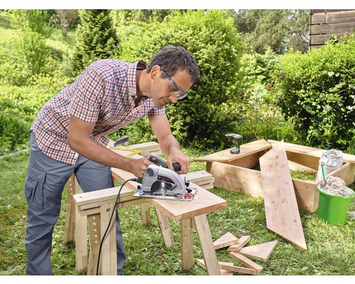 Ein Mann sägt Holz mit einer Handkreissäge im Garten. Werkzeuge und Baumaterialien sind im Hintergrund zu sehen.