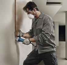 Handwerker arbeitet mit einer Mauernutfräse an einer Wand. Er trägt Schutzbrille, Mundschutz und Handschuhe.