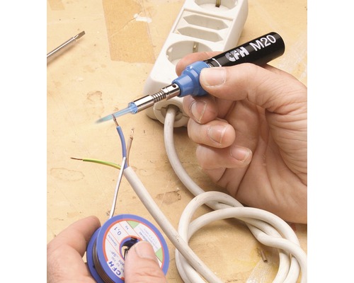 Eine Hand lötet eine elektrische Leitung mit einem Lötkolben. Auf dem Tisch liegen eine Spule Lötzinn und eine Steckdosenleiste.