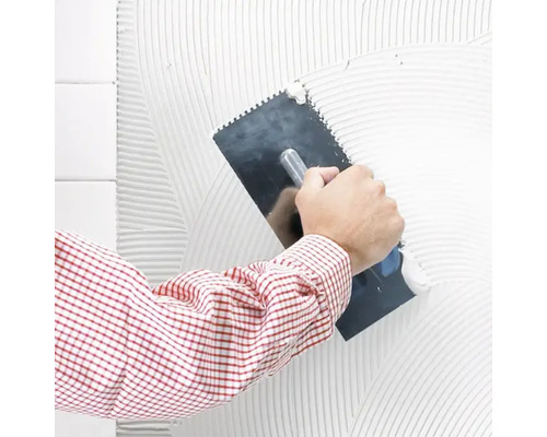 Ein Mann trägt mit einer Zahnspachtel Fliesenkleber auf eine Wand auf.