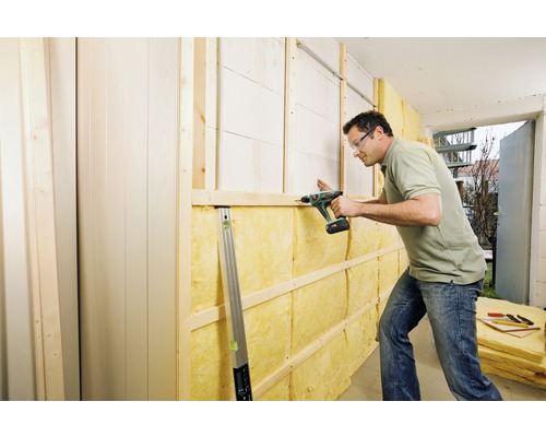 Ein Mann befestigt Dämmwolle an einer Wand mit einem Akkuschrauber. Holzrahmen und Paneele sind sichtbar.