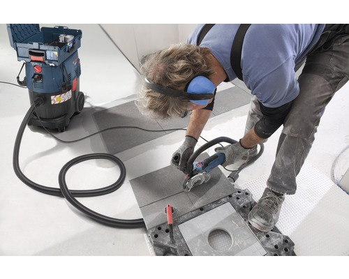 Ein Handwerker schneidet eine Fliese mit einem Winkelschleifer, wobei ein Industriesauger Staub absaugt.