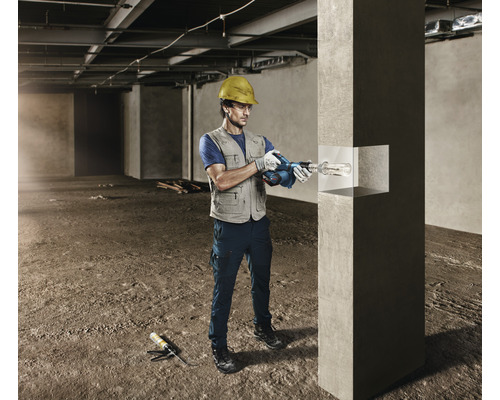 Ein Mann mit Schutzhelm bohrt mit einem Bohrhammer in einen Betonpfeiler auf einer Baustelle.