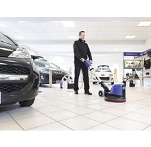 Mann reinigt einen Ausstellungsraum mit einer Reinigungsmaschine, Autos sind im Hintergrund zu sehen.